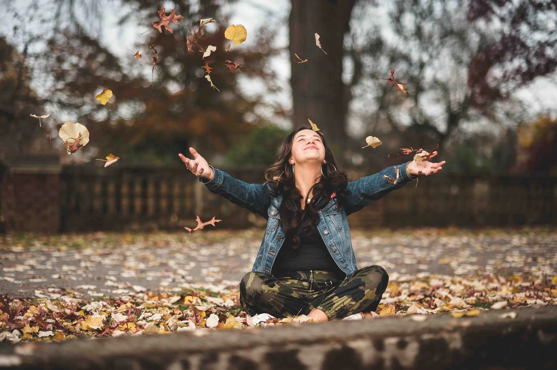 Alexis tossing autumn leaves into the air while sitting cross-legged.