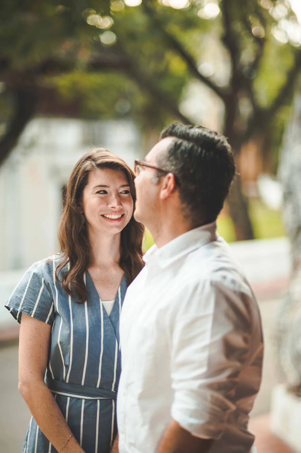 A couple laughing together with trees and a white railing behind them.
